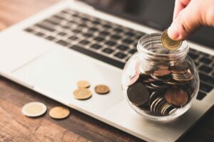 כמה עולה פרסום ממומן בגוגל. כף יד מכניסה מטבע לצנצנת מלאה מטבעות שיושבת על מקלדת לפטופ.